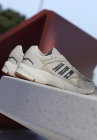 Jasno beżowe buty sportowe z siateczkową i skórzaną teksturą, z trzema srebrnymi paskami, białą podeszwą środkową i podeszwą z gumy.
