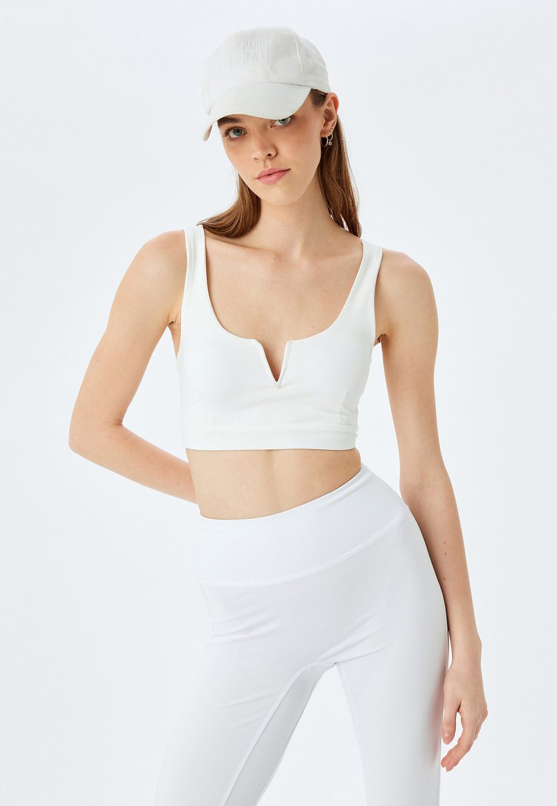 Sujetador deportivo blanco recortado con escote en V. Combinado con leggings blancos de cintura alta. El modelo lleva una gorra de color claro. Tejido suave y elástico.