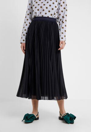 Kobieta ubrana w czarną plisowaną spódnicę midi, białą bluzkę w brązowe grochy oraz zielone płaskie buty z dużymi kokardami i perłowymi paskami.