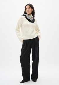 Kvinde står med hænderne i lommerne iført en cremegul kabelstrik sweater, sorte vide bukser, spidse sko og et stribet sort-hvidt tørklæde.