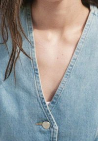 Blåt denimtøj med V-hals, der har en sølvfarvet knaplukning og synlige syningsdetaljer. Blød tekstur, afslappet pasform.