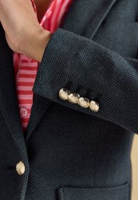 Manica di blazer scuro a motivo spina di pesce con quattro bottoni metallici decorativi, indossato sopra una camicia a righe rosse e rosa.