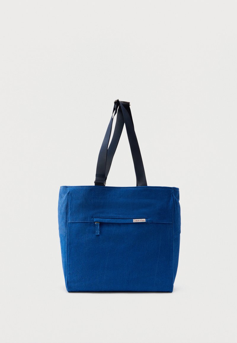 Sac fourre-tout en toile bleue avec une poche zippée à l'avant, des doubles bretelles et une forme rectangulaire. Dispose d'une étiquette de marque discrète.
