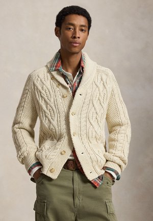 Jeune homme portant un cardigan en maille torsadée crème sur une chemise à carreaux, un pantalon cargo vert et les mains dans les poches sur un fond neutre.