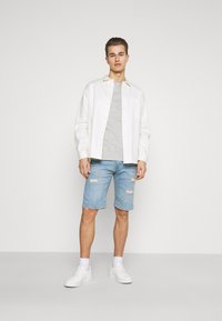 Hvit skjorte over en grå t-skjorte, kombinert med lyseblå, slitte denimshorts og hvite joggesko, stående mot en enkel bakgrunn.