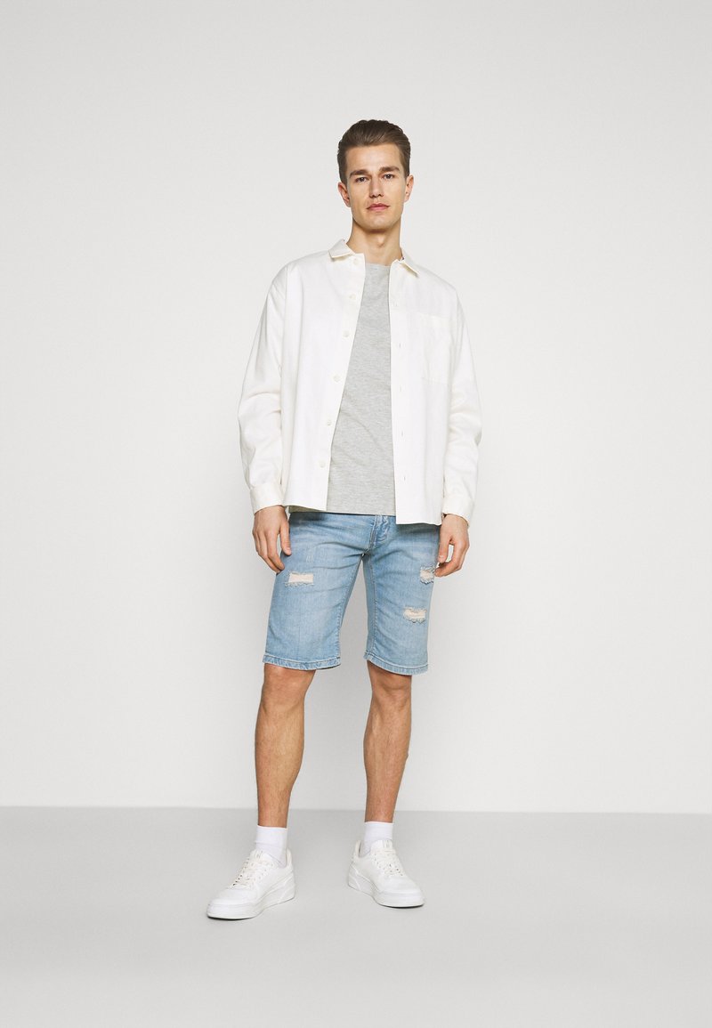 Hvit skjorte over en grå t-skjorte, kombinert med lyseblå, slitte denimshorts og hvite joggesko, stående mot en enkel bakgrunn.