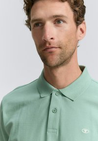 Mintgrøn polo T-shirt med krave, to knapper og broderet logo på venstre side. Stoftekstueren virker blød og åndbar.