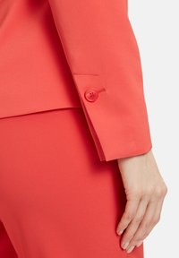 Korálový blazer s rukávy v přiléhavém střihu, s kulatým červeným knoflíkem, hladkou texturou a ohnutým detaily na manžetách.