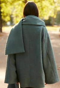 Hrstkově zelený kabát s měkkou texturou, volným střihem a širokými rukávy. Obsahuje velký, splývavý límec. Pohled zezadu.