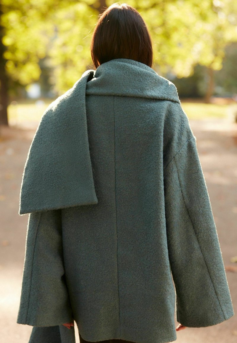 Hrstkově zelený kabát s měkkou texturou, volným střihem a širokými rukávy. Obsahuje velký, splývavý límec. Pohled zezadu.