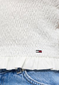 Λευκό τοπ από ρούχα με ρίγες και μικρό λογότυπο Tommy Hilfiger πάνω από το βολάν στο τελείωμα, σε συνδυασμό με μπλε τζιν τζιν που είναι ορατά στη μέση.