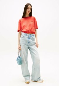 Kvinde iført en koralfarvet cropped T-shirt, lyseblå brede jeans, hvide sneakers og holder en lille blå håndtaske med kædestrop.