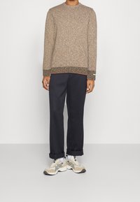 Brauner strukturierter Pullover mit rundem Halsausschnitt und kontrastierendem Saum, kombiniert mit dunklen hochgekrempelten Hosen und hellen Sneakern mit schwarzen Akzenten.