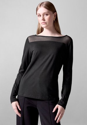 Black long-sleeve top with a sheer mesh panel at the neckline, smooth texture, and a relaxed fit. Paired with black pants.