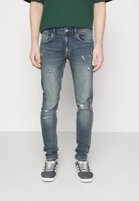 Jeans de denim azul desgastado con detalles rasgados, corte ajustado, diseño de cinco bolsillos y cierre de botón. Llevados con zapatillas grises y camisa verde.