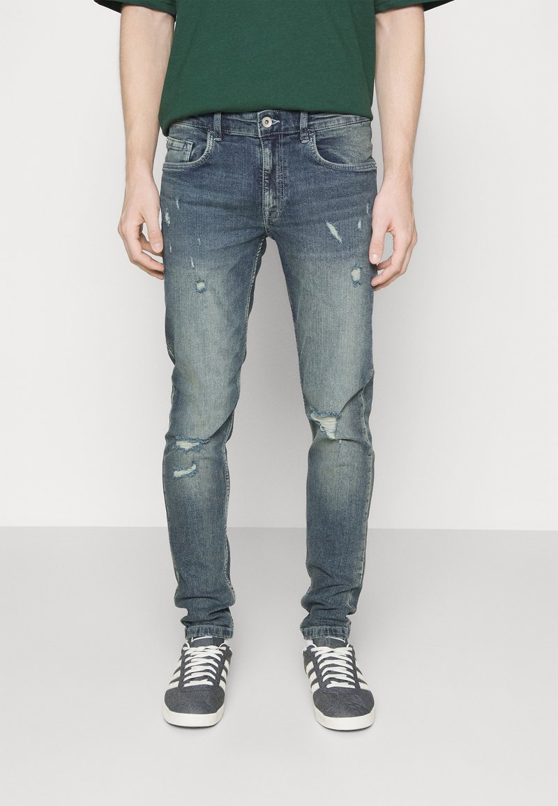Jeans de denim azul desgastado con detalles rasgados, corte ajustado, diseño de cinco bolsillos y cierre de botón. Llevados con zapatillas grises y camisa verde.