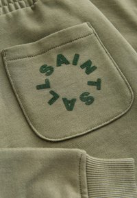 Olivenfarbener Stoff mit einer Tasche, die mit kreisförmigem, dunkelgrünem, gesticktem Text "SAINT" versehen ist. Die Nähte sind ordentlich mit gerippten Details.