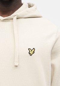 Felpa con cappuccio color beige con finitura strutturata, con logo aquila ricamato in nero e oro sul petto a sinistra. Cappuccio con coulisse incluso.