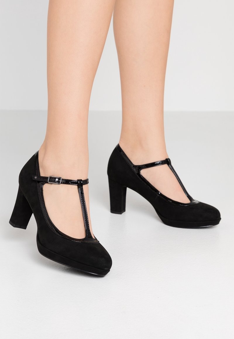 Escarpins à talons hauts en daim noir avec bride en T et bouts arrondis, portés pieds nus sur un fond clair uni.