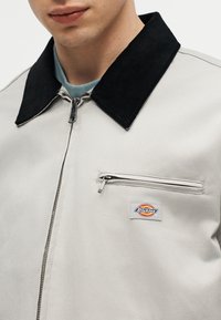 Gros plan sur une personne portant une veste zippée gris clair avec un col noir, une poche zippée sur la poitrine et un patch logo Dickies sur la poche.
