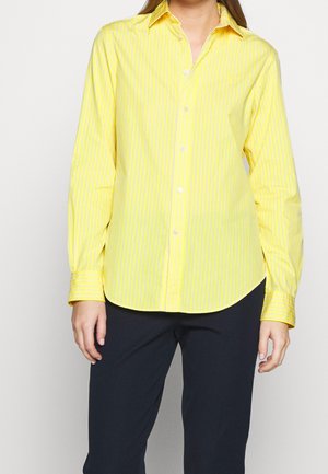 Femme portant une chemise jaune à manches longues à rayures verticales claires et un pantalon noir, debout devant un fond uni.