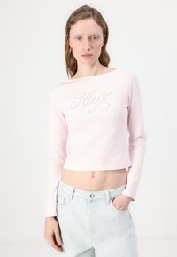 Šviesiai rožinė trumpa ilgomis rankovėmis palaidinė su lokio logotipu, išklotu blizgučiais krūtinės srityje, pagaminta iš minkštos medžiagos. Dėvima su šviesiai mėlynomis džinsomis.
