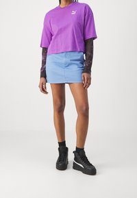 Fialové zkrácené tričko s bílým logem, světle modrá denimová mini sukně a černé vysoké tenisky s bílými akcenty a texturovaným povrchem.