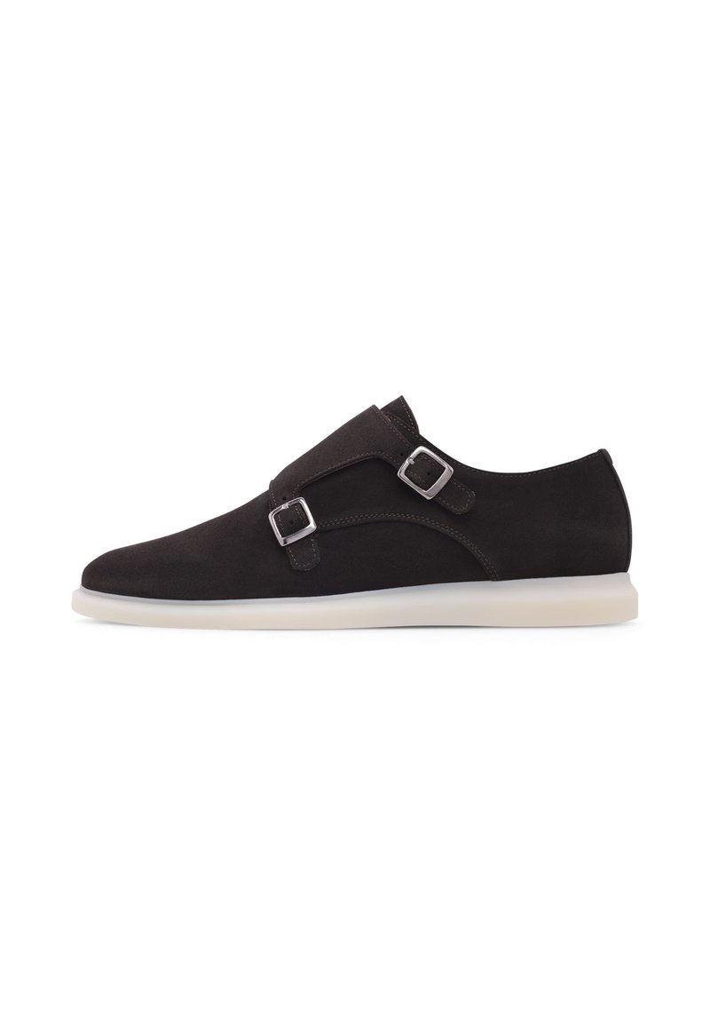 Chaussure slip-on en suede marron foncé avec deux boucles argentées, un bout arrondi et une semelle en caoutchouc blanche contrastante. Texture lisse, design minimal.