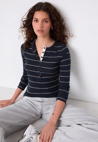 Haut à manches longues à rayures bleu marine et blanches avec un col Henley, comprenant cinq boutons et une texture côtelée. Associé à un pantalon de survêtement gris.