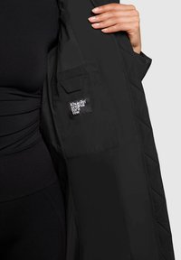 Een persoon in zwarte kleding houdt een zwarte gewatteerde jas open en toont de binnenzak met het label "Khujo original for a few".