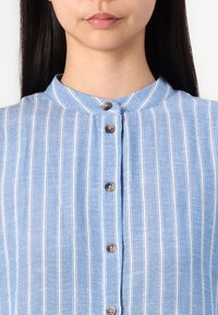Camisa azul clara com riscas verticais brancas, um fecho frontal com botões castanhos e um colarinho redondo. Tecido texturizado.