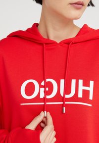 Punainen hupullinen collegepaita, valmistettu pehmeästä kankaasta, jossa on näkyvä valkoinen logo, säädettävät nyörit hopeanvärisillä päillä ja resorit hihansuissa.