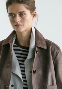 Brun læderjakke med en bred krave lagdelt over en grå og hvidstribet sweater. Jakken har knapdetaljer og en struktureret overflade.