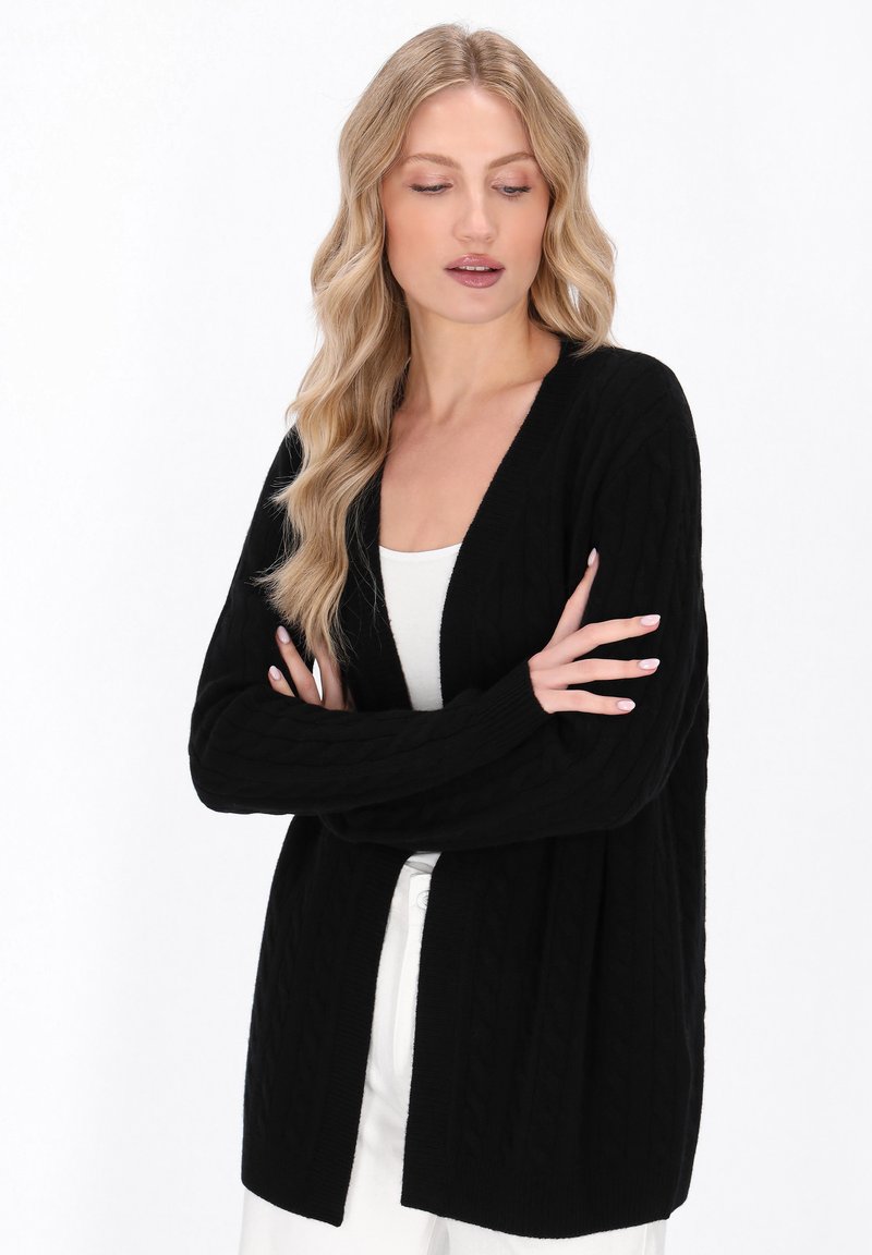 Cardigan nera a lavorazione a trecce, con maniche lunghe, apertura frontale e bordi a costine, realizzato in un tessuto morbido, abbinato a una maglia bianca e pantaloni chiari.
