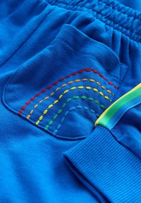 Pantaloni scurți din bumbac albastru, cu o buzunar în spate decorat cu un detaliu curcubeu cusut multicolor. Talie elastică și mansete texturate.