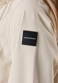 Giacca beige con una texture liscia, caratterizzata da una toppa rettangolare nera con la scritta "SWEDEMOUNT" in bianco in rilievo. Dettagli delle cuciture visibili sulle maniche.