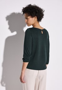 Haut vert foncé à manches trois quarts avec un col rond et un détail en trou de serrure à l'arrière, associé à un pantalon de couleur claire.