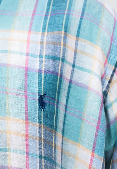 Detailný záber na modro-tyrkysovo-viacfarebnú károvanú látku s malým námornícky modrým vyšívaným logom koňa a jazdca.