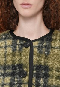 Κοντινό πλάνο στο κάτω μέρος του προσώπου και τον λαιμό μιας γυναίκας που φοράει ένα κουμπωτό, υφασμάτινο μάλλινο ζακέτα σε πράσινο και μαύρο με μαύρο περίγραμμα.