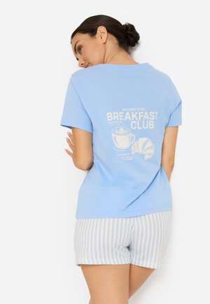 Γυναίκα με φωτεινό γαλάζιο t-shirt με κείμενο "Breakfast Club", κούπα καφέ και γραφικό κρουασάν, σε συνδυασμό με λευκό και μπλε ριγέ σορτς.