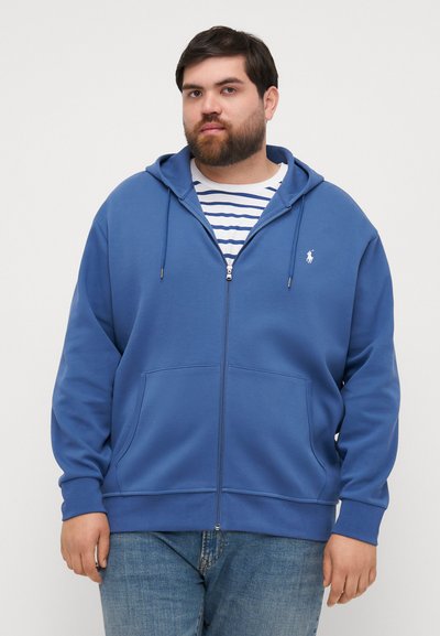Plava zip-hoodie od mekog materijala s dva prednja džepa, kapuljačom s vezicom i malim logom na prsima, nosi se preko prugaste košulje.