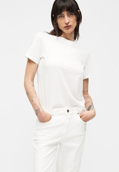 Γυναίκα με σκουρόχρωμα μαλλιά που φοράει απλό λευκό t-shirt και λευκό παντελόνι, στέκεται με τα χέρια στις τσέπες, δείχνοντας πολλά τατουάζ και στα δύο μπράτσα.