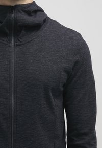Zwarte hoodie met ritssluiting, gemaakt van een gestructureerde stof. Heeft een hoge kraag en een aansluitend ontwerp. Naaddetails zijn zichtbaar aan de voorkant.