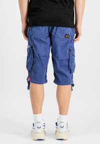 Zili cargo šorti ar vairākiem kabatām, dekoratīvām šuvēm un regulējamiem apakšmalām, saskaņoti ar baltiem zeķēm un sporta apaviem.