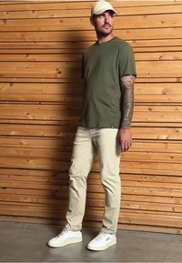 Camiseta de manga corta verde oliva, pantalones beige ajustados, zapatillas blancas con detalles verdes, de pie contra un fondo de pared de madera.