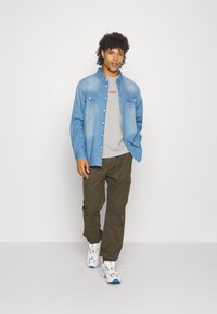 Camisa de mezclilla con dos bolsillos en el pecho, camiseta gris con logo rojo, pantalones cargo oliva y zapatillas blancas con detalles en azul.