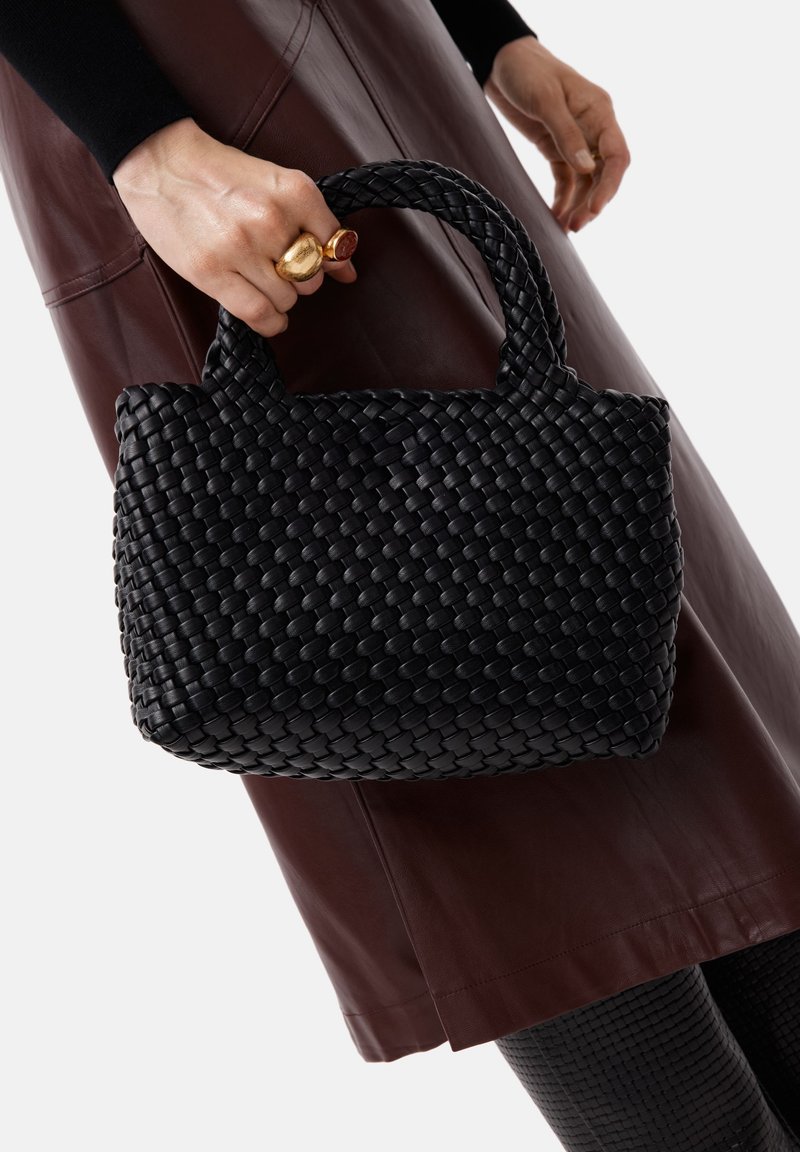 En hand håller en svart flätad handväska, bärande en brun läderskjol, svart topp, ringar och texturerade svarta leggings mot en vit bakgrund.