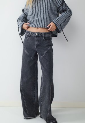 Person trägt weit geschnittene, ausgewaschene schwarze Jeans mit sichtbaren Nahtdetails und einen grauen Rippenpullover, Hände justieren den Pullover-Saum.