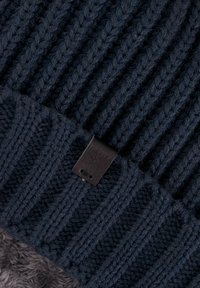 Navy blauwe gebreide beanie met geribbelde textuur, voorzien van een zwart leren label aan de onderkant. Zacht, warm, met een opvouwbaar ontwerp.