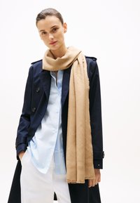 Beige tørklæde lavet af blødt materiale, med et vævet mønster og frynsede kanter, båret over en lyseblå skjorte og en navy trenchcoat.